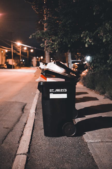 A group of tightly packed black plastic rubbish bags, placed on the edge of a sidewalk next to a wooden fence and a bush with dense foliage. The bags appear to contain various types of waste and are p