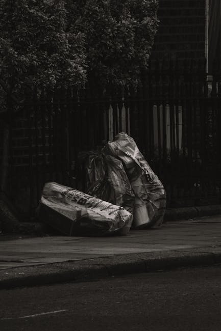 A narrow urban alleyway cluttered with a large, black wheeled waste container in the foreground, surrounded by scattered rubbish including paper and plastic debris on the uneven asphalt ground. In the