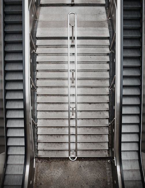 A straight, upward-facing view of a modern outdoor staircase constructed from dark, polished stone or concrete, featuring wide, evenly spaced steps with a smooth, reflective surface. The staircase has
